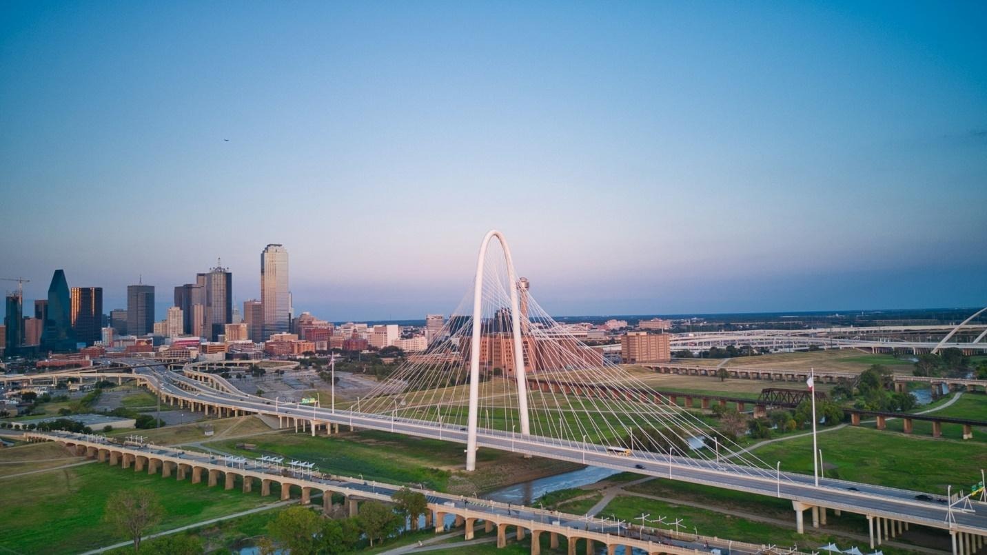 Aerial view of a city skyline at sunset with a prominent cable-stayed bridge in the foreground. The sky is clear, creating a serene atmosphere.